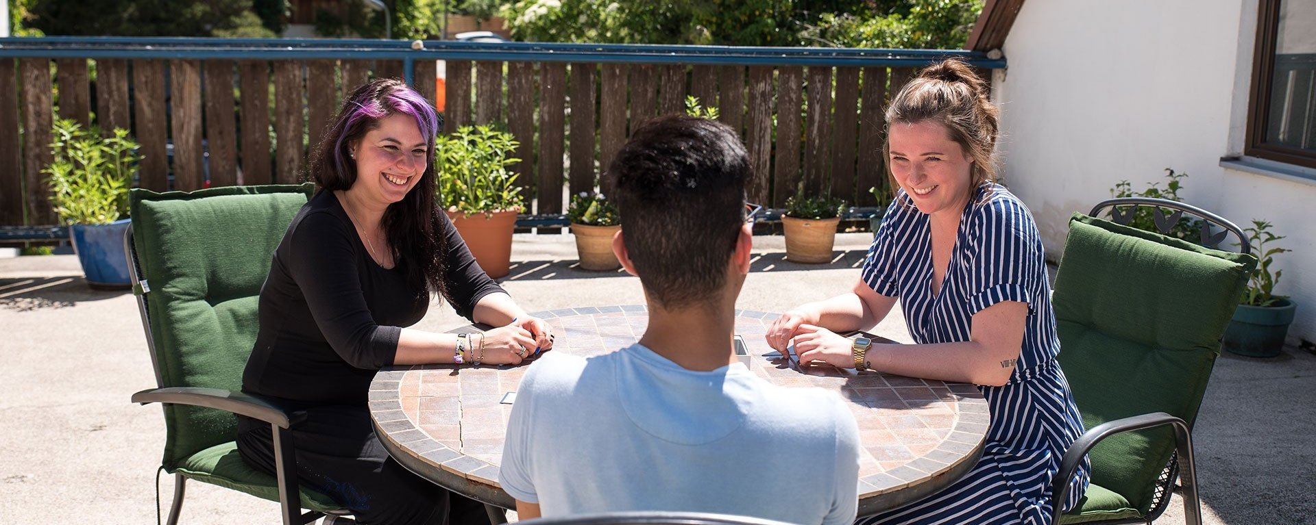 Zwei junge Frauen und ein Mann sitzen an einem steinernen Gartentisch - Der Mann scheint einen Spaß gemacht zu haben - beide Frauen lachen