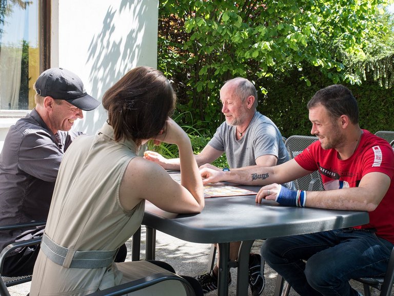 eine Frau und drei Männer sitzen am Gartentisch und spielen ein Brettspiel