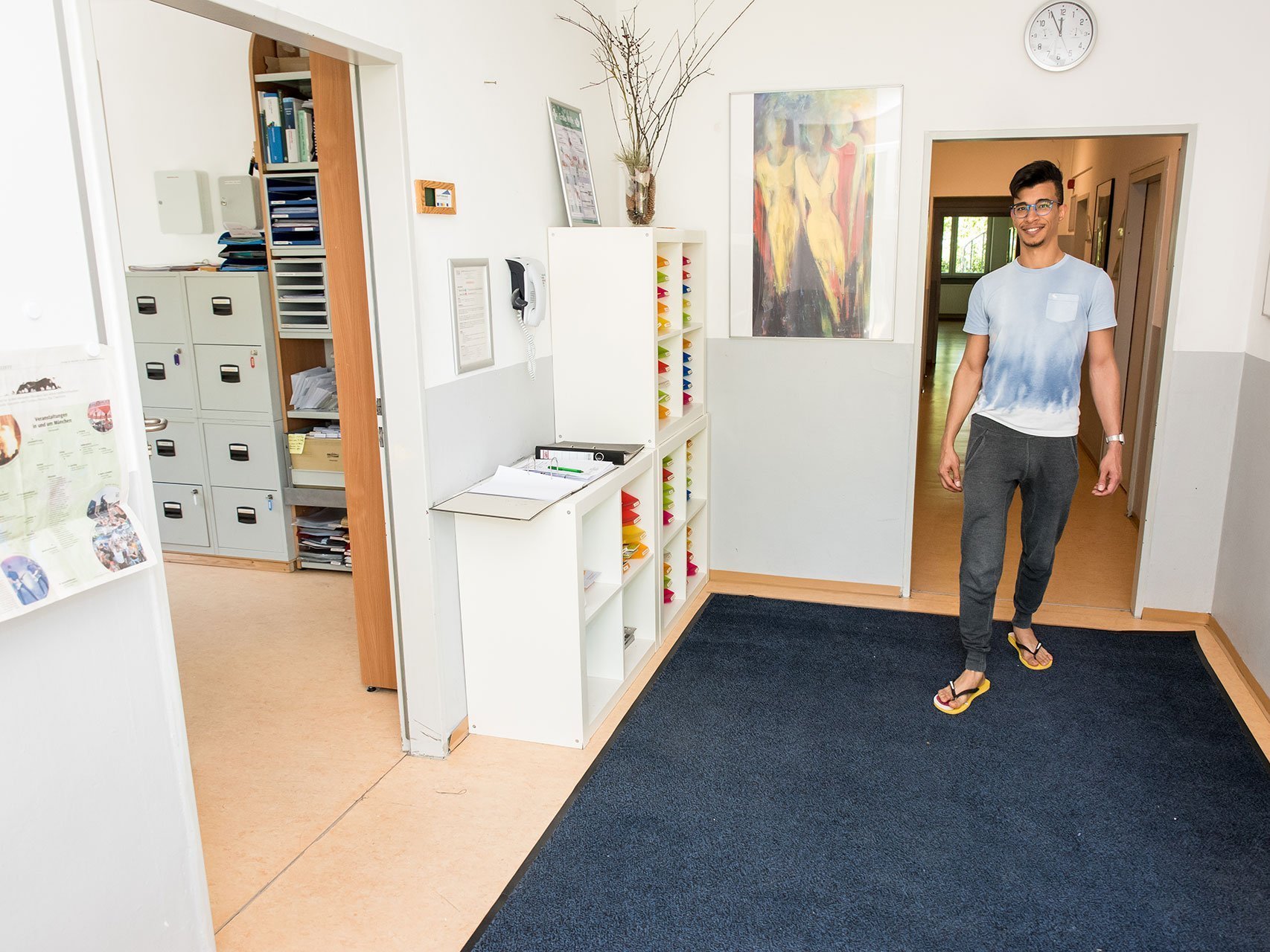 Ein junger Mann mit Brille, Jogginghose und Flip-Flops betritt lächelnd ein Büro